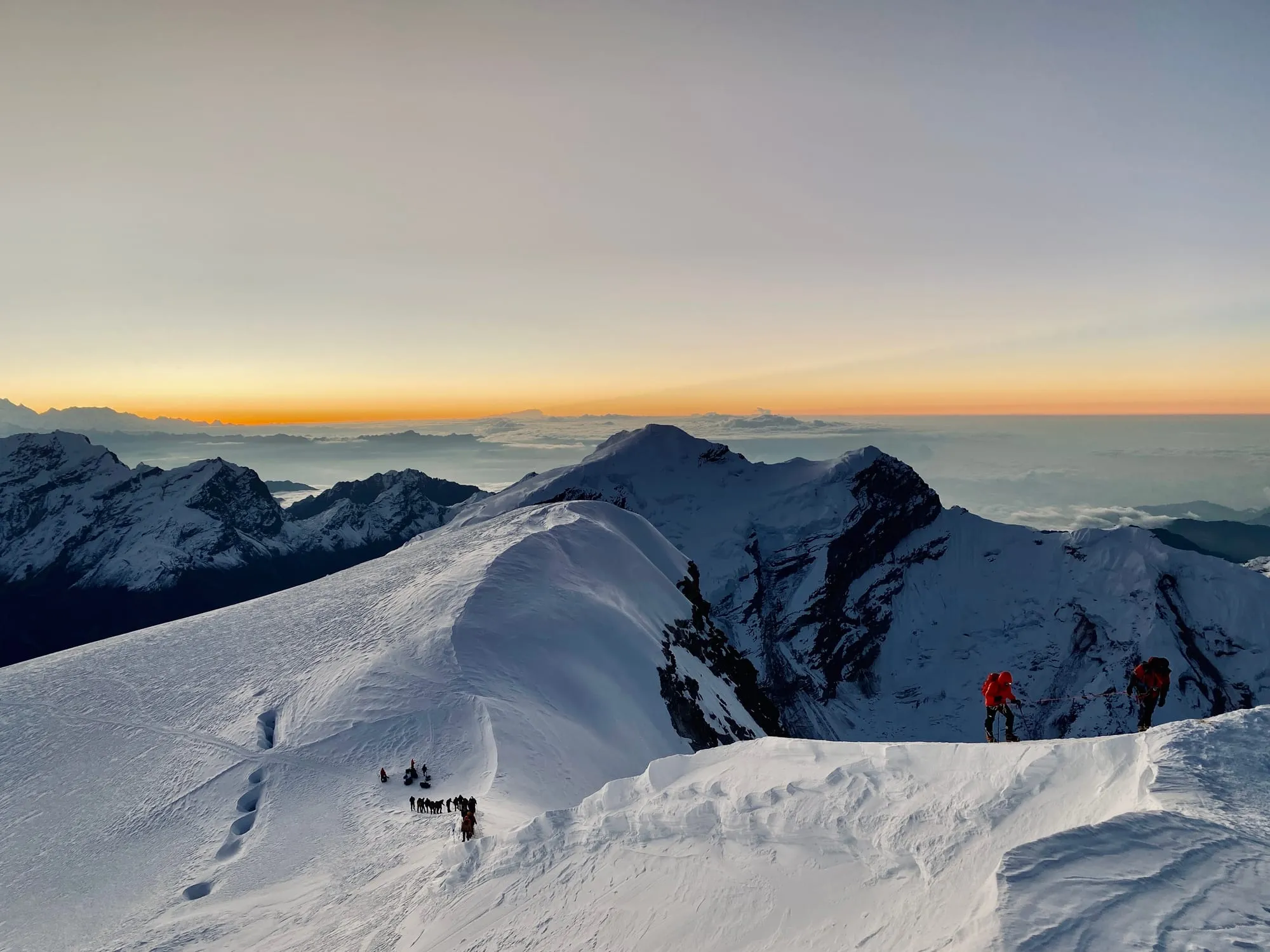 Climbers strung out along a broad snow ridge at first light, a sea of cloud below them and the sky turning orange behind the peaks.