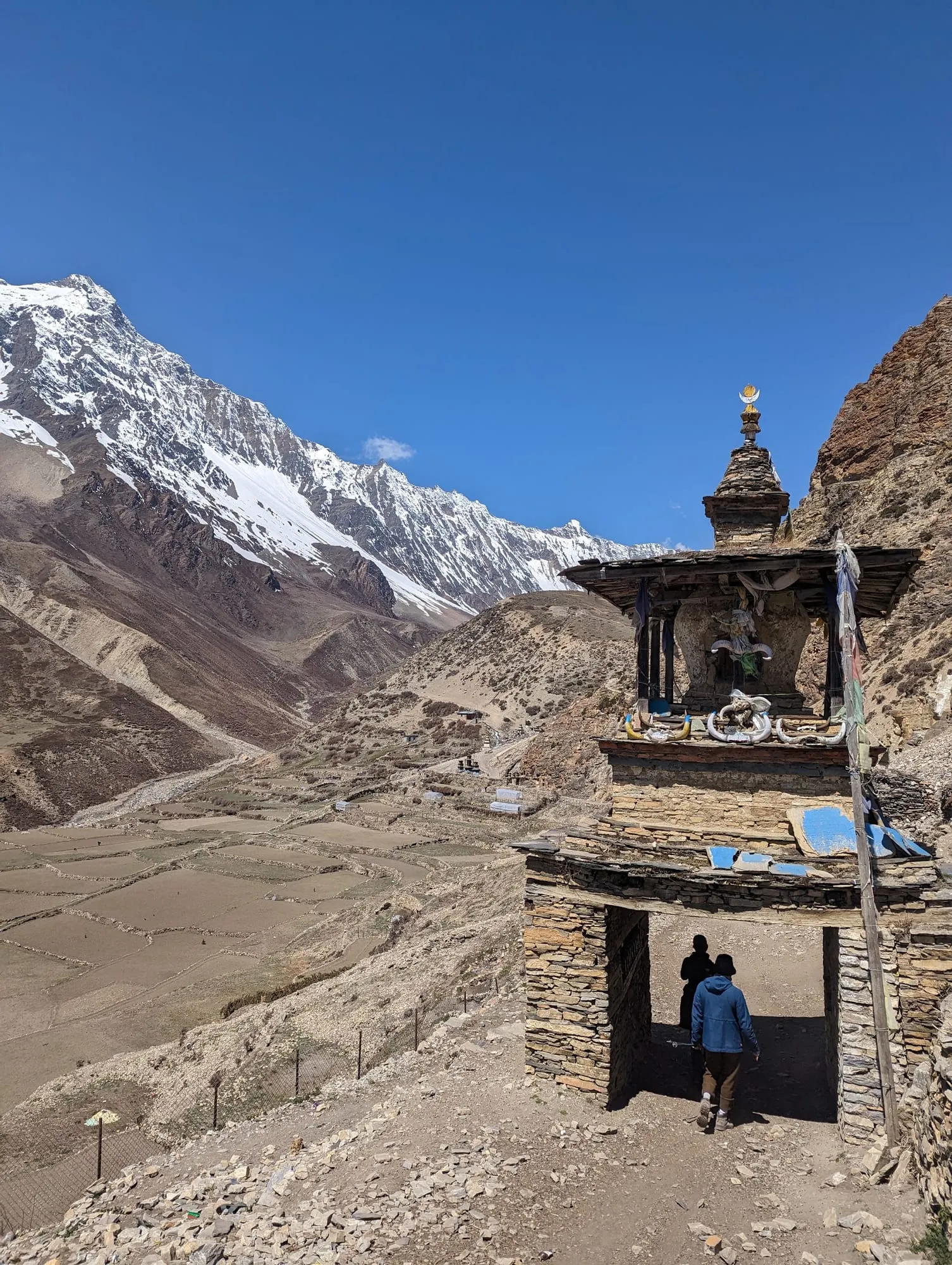 A trekker passing under a stone kani gateway topped with a chorten, the dry valley and its snow peaks opening up beyond.