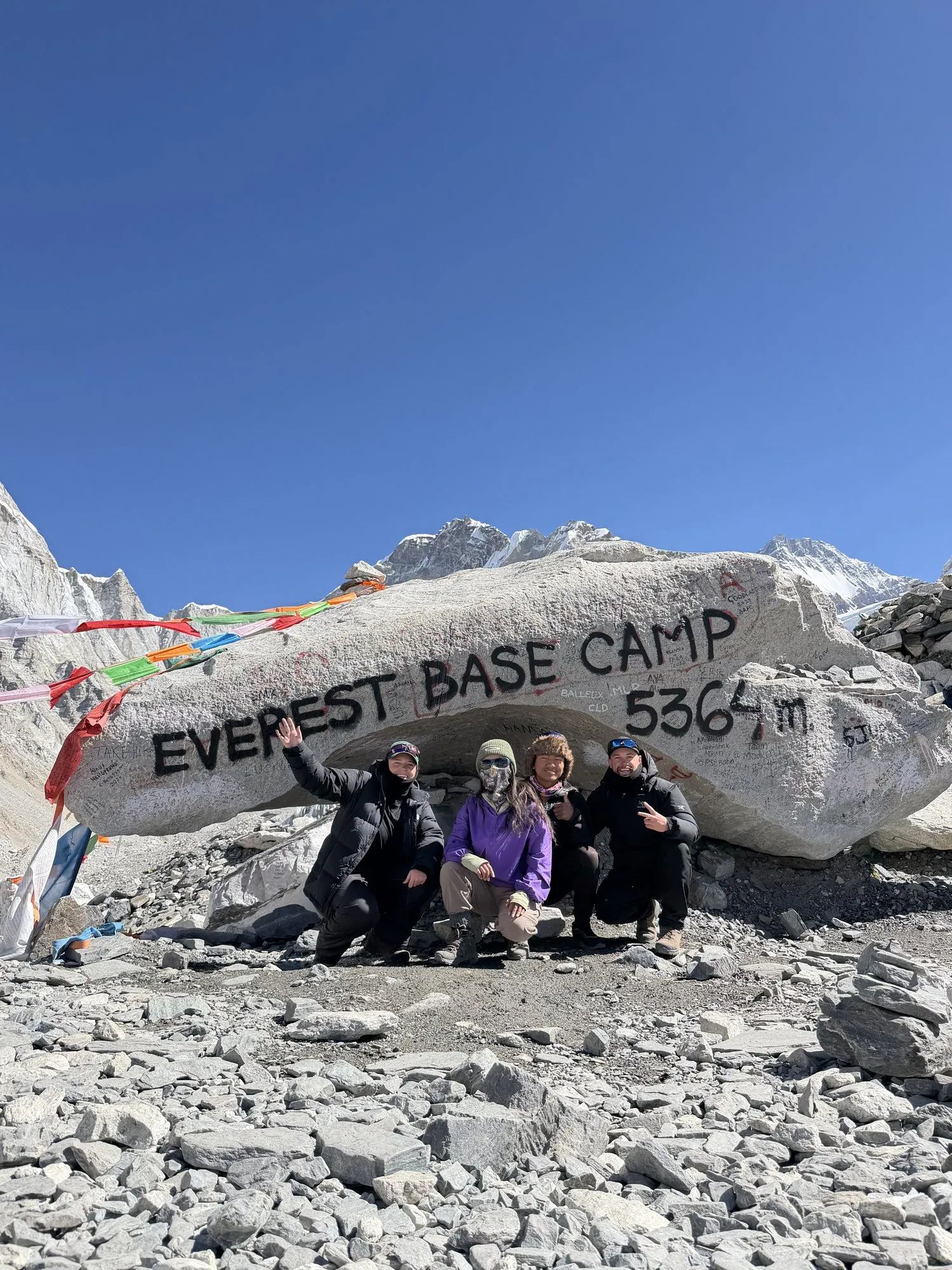 Trekkers sitting together at the Everest Base Camp boulder, hung with prayer flags and painted with the altitude, 5,364 m, under a hard blue sky.