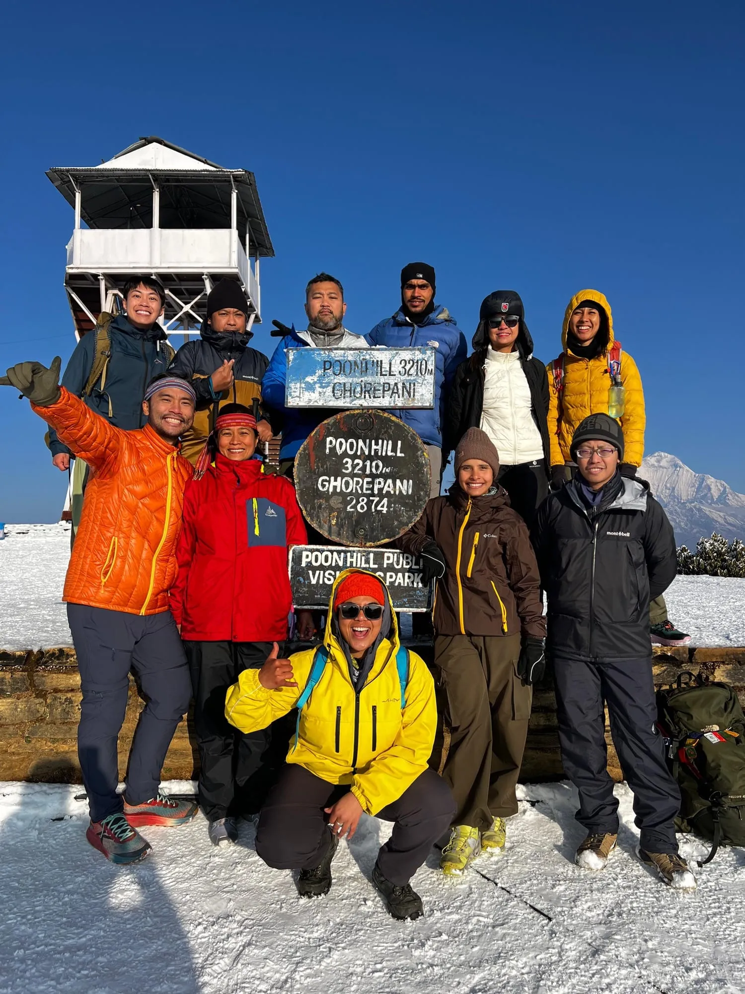 A group of trekkers and their guides gathered around the Poon Hill marker stone reading 3,210 m, snow underfoot and the viewing tower behind them.