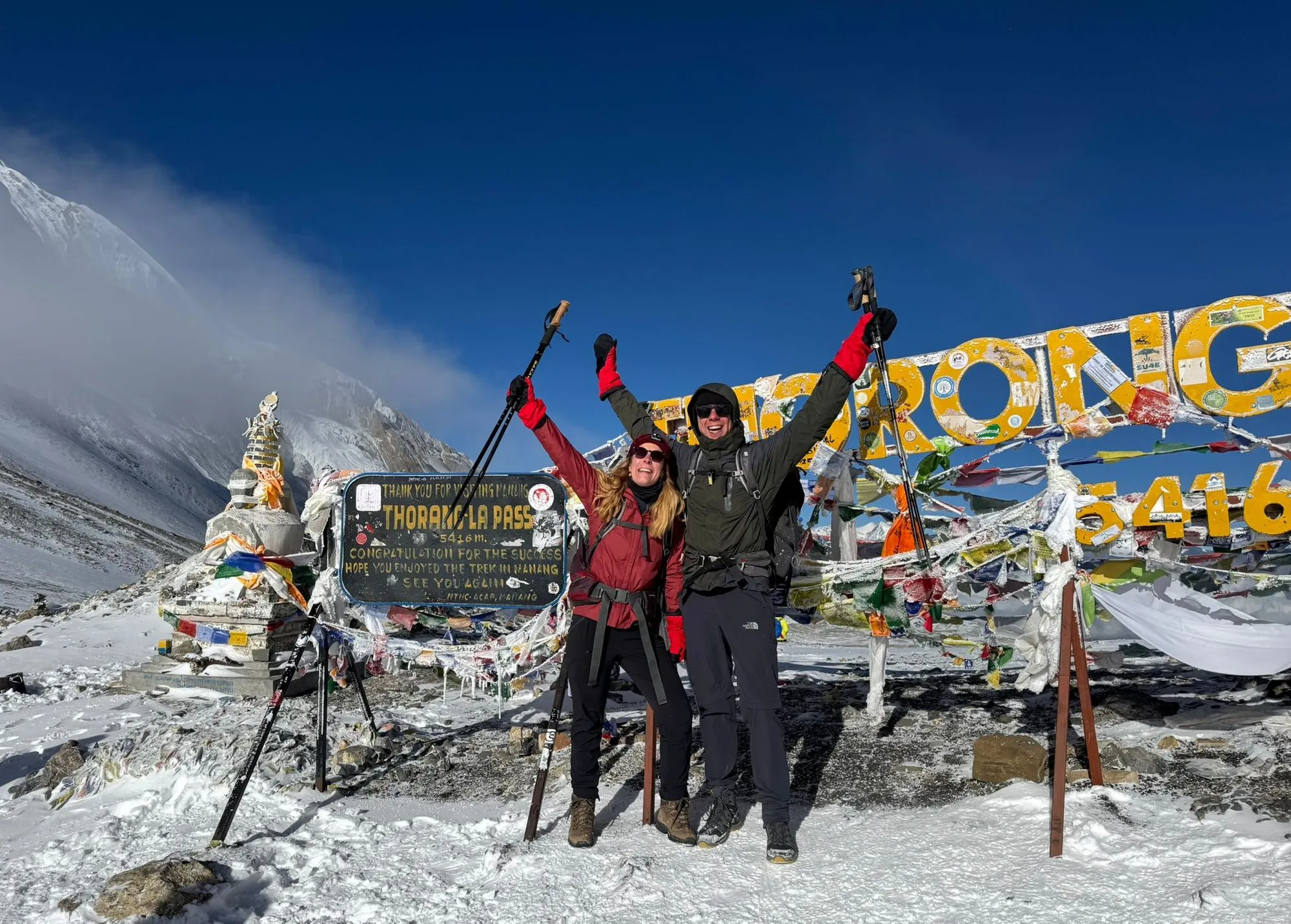 Two trekkers on the snow at the Thorong La, poles raised, beside the painted pass sign and its lines of prayer flags.