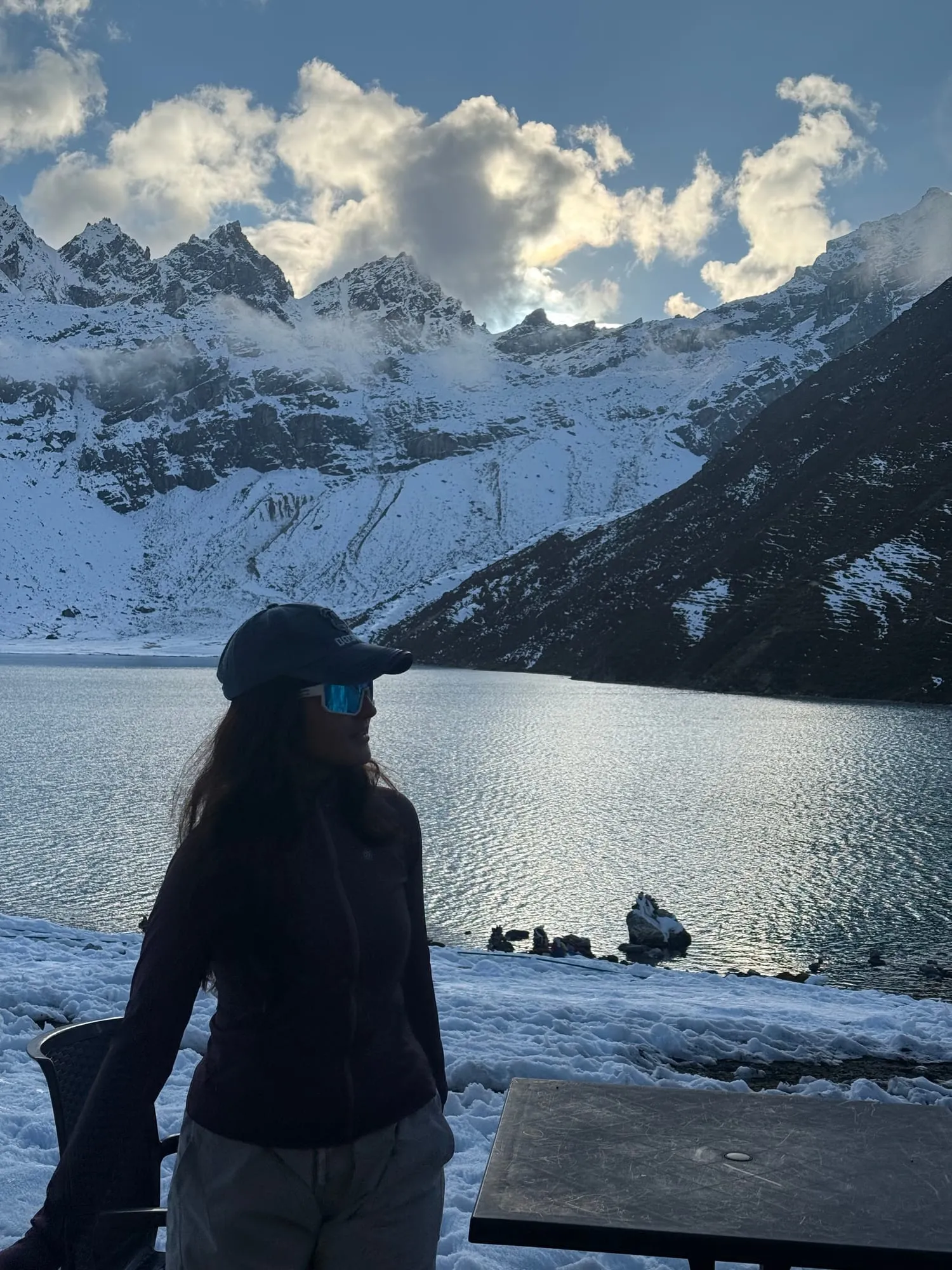 A trekker in a cap and sunglasses standing on the snowy shore of a Gokyo lake, the water bright against dark rock and snow-covered peaks behind.