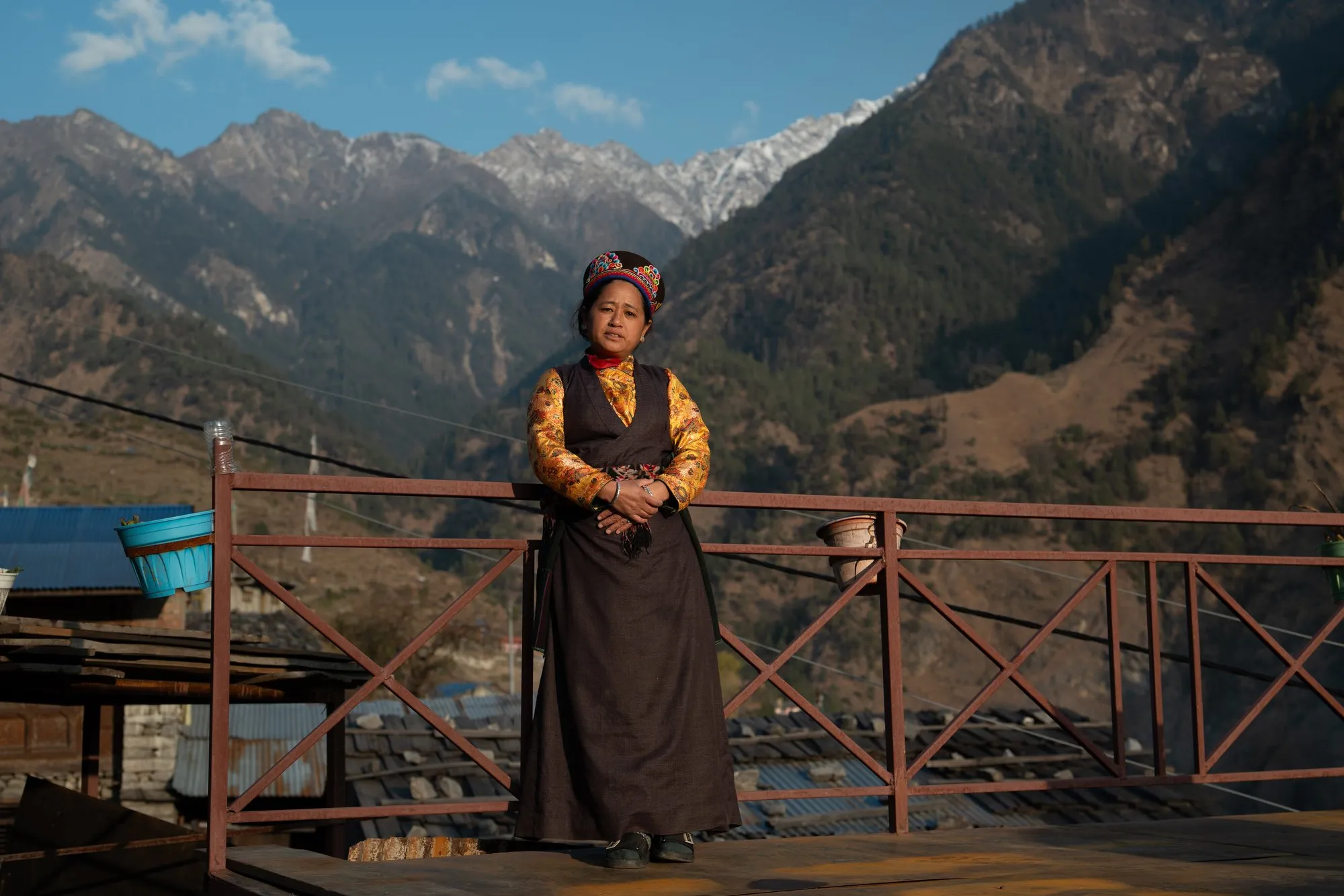 A woman in traditional Tamang dress standing at a wooden balcony rail, the forested Langtang valley and its ridgelines opening out behind her.