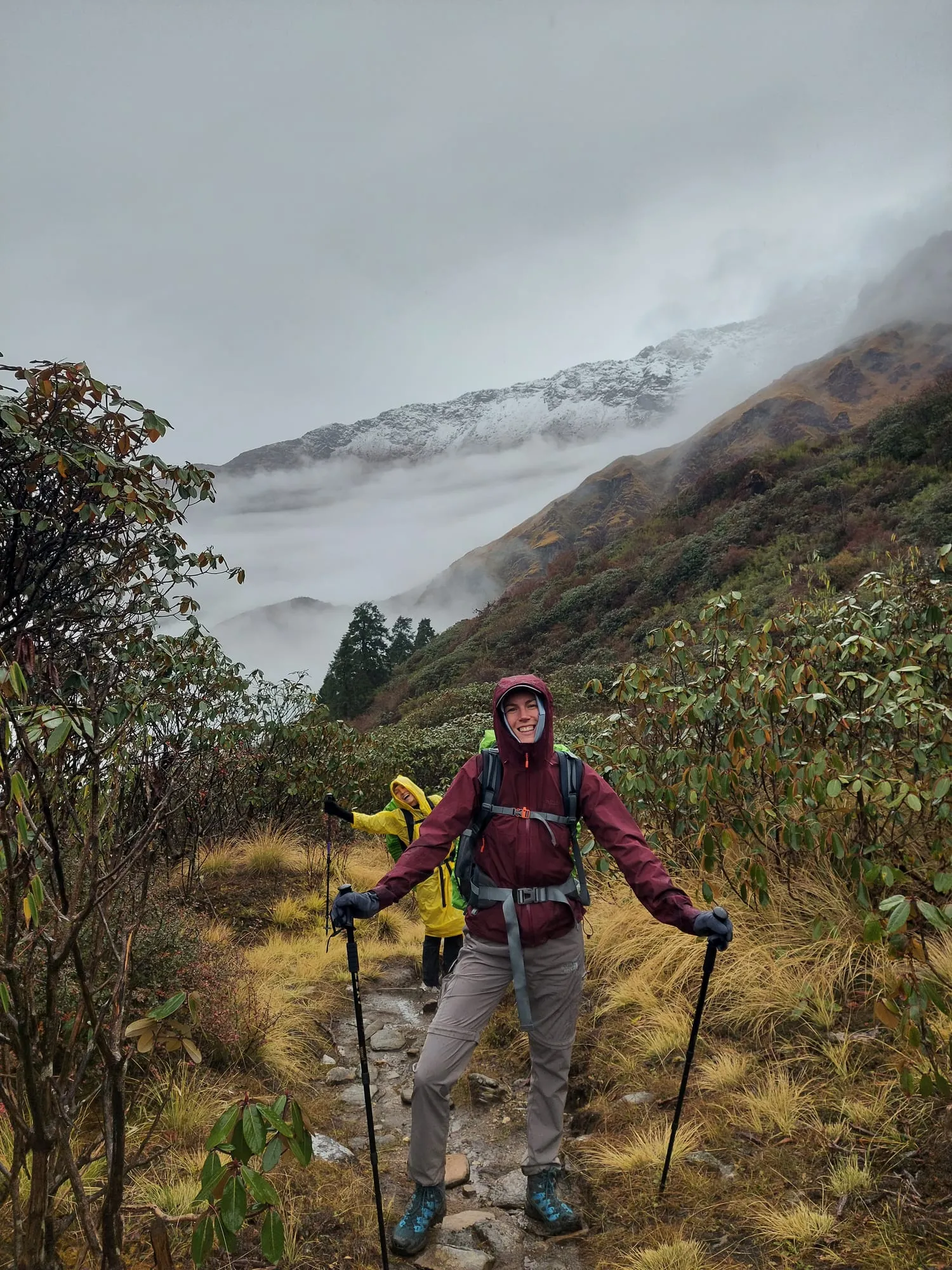 A trekker in a maroon rain jacket standing on the wet trail with poles, a second walker behind in yellow, cloud filling the valley and fresh snow on the ridge above.