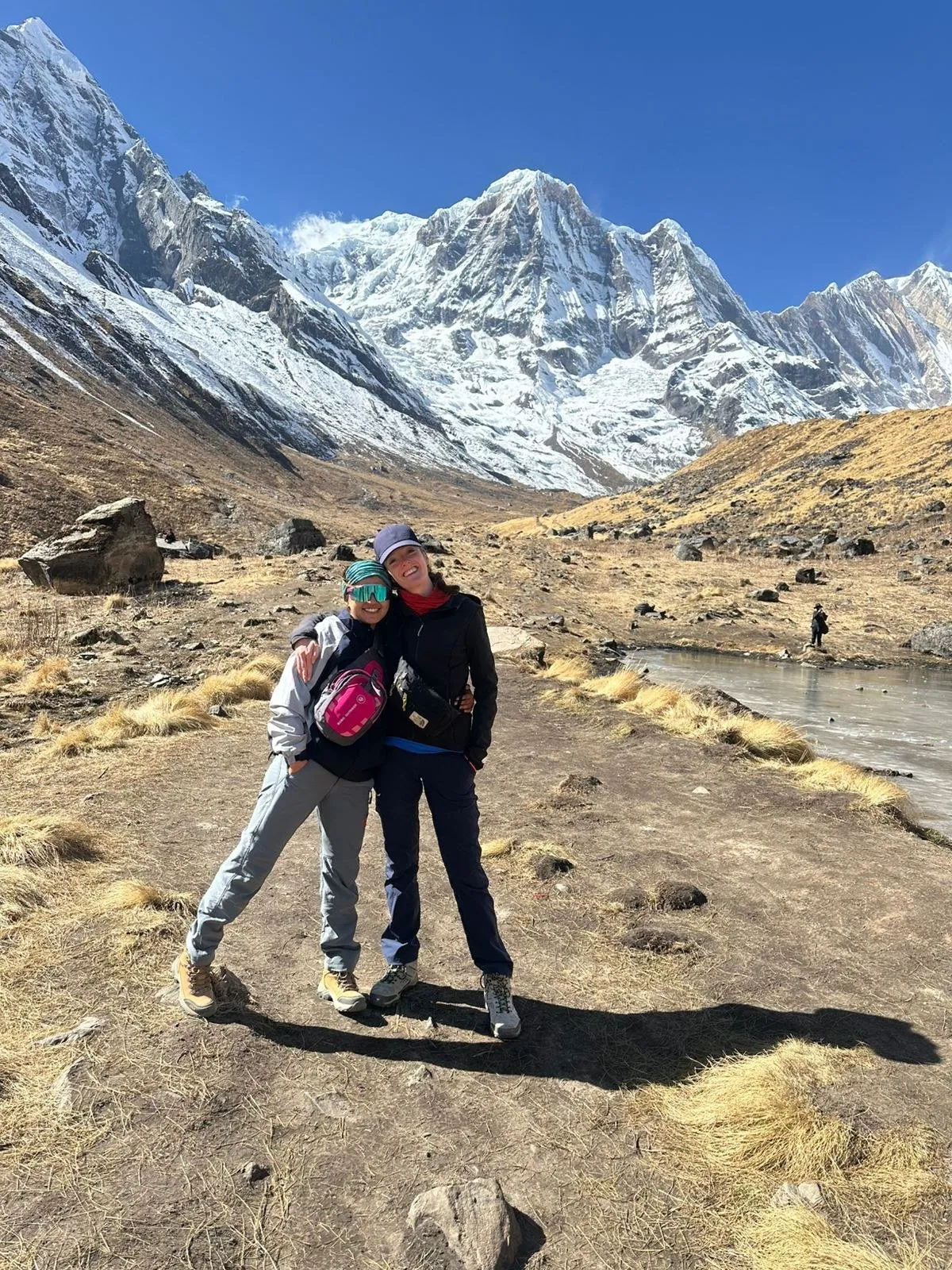 Guide Ashmita Poudel and a traveller with their arms round each other on the floor of the Annapurna Sanctuary, snow-covered peaks rising directly behind them.