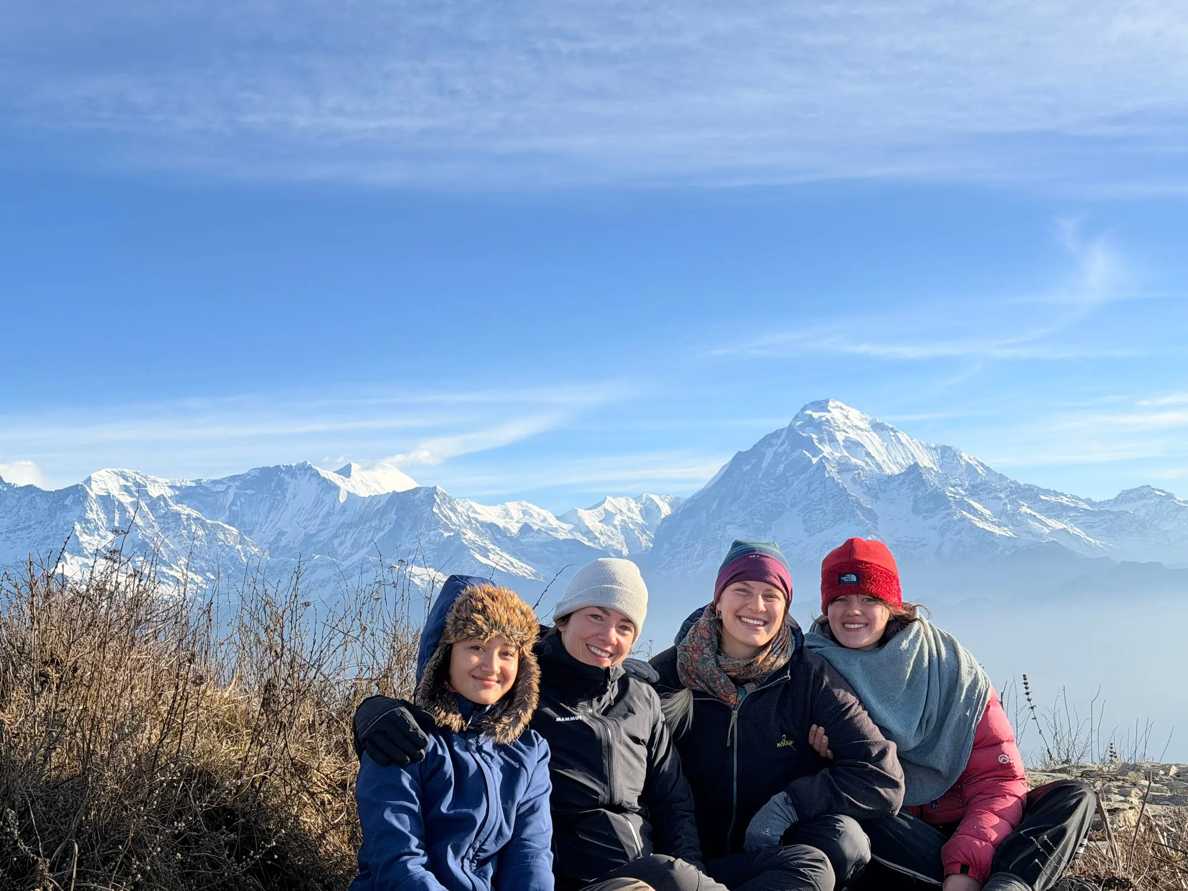 Four trekkers sitting close together on a ridge in the early light, the Annapurna peaks rising above a sea of cloud behind them.
