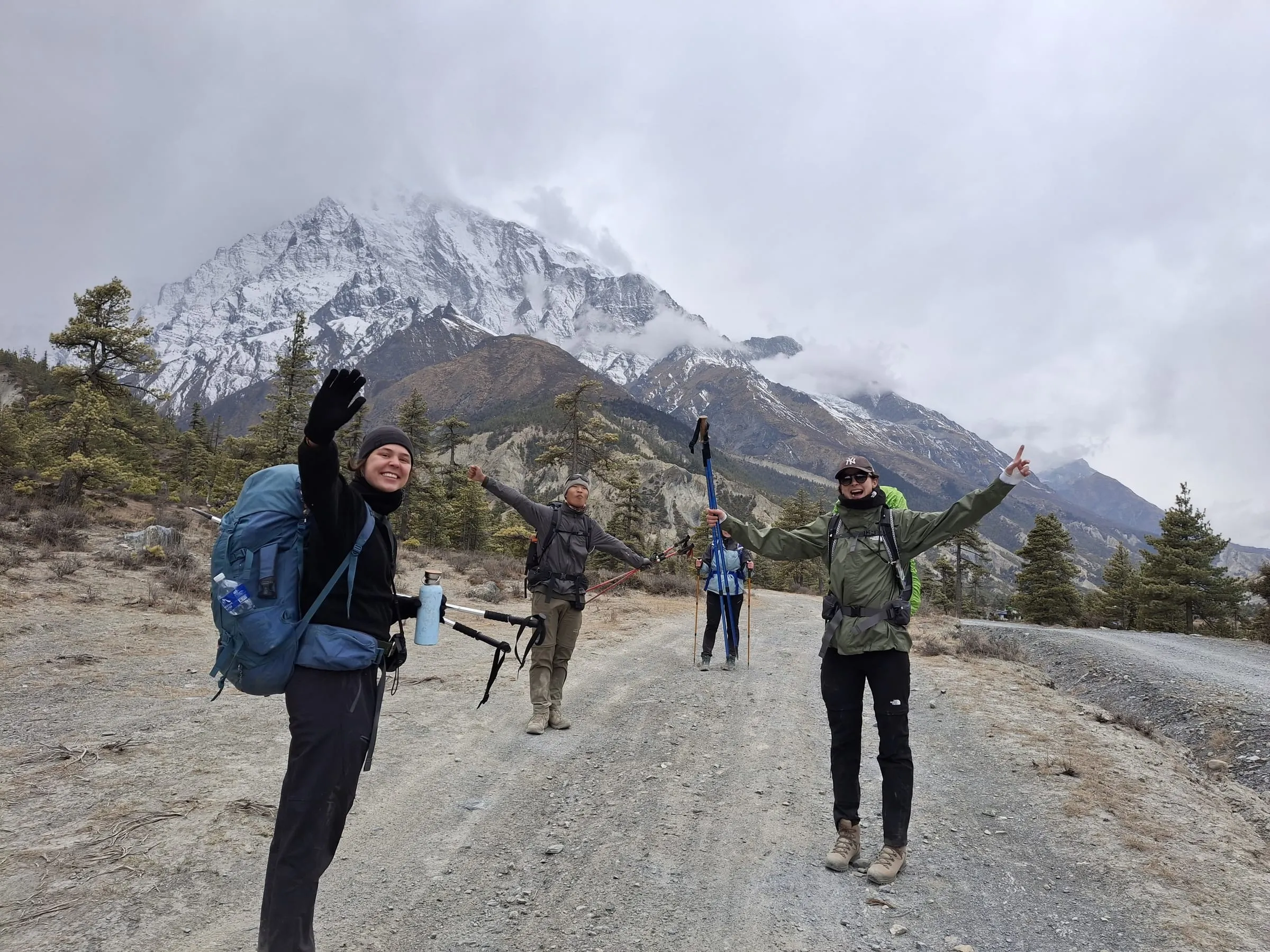 Three trekkers with rucksacks and poles raised on a gravel mountain road, a cloud-covered snow peak filling the valley behind them.