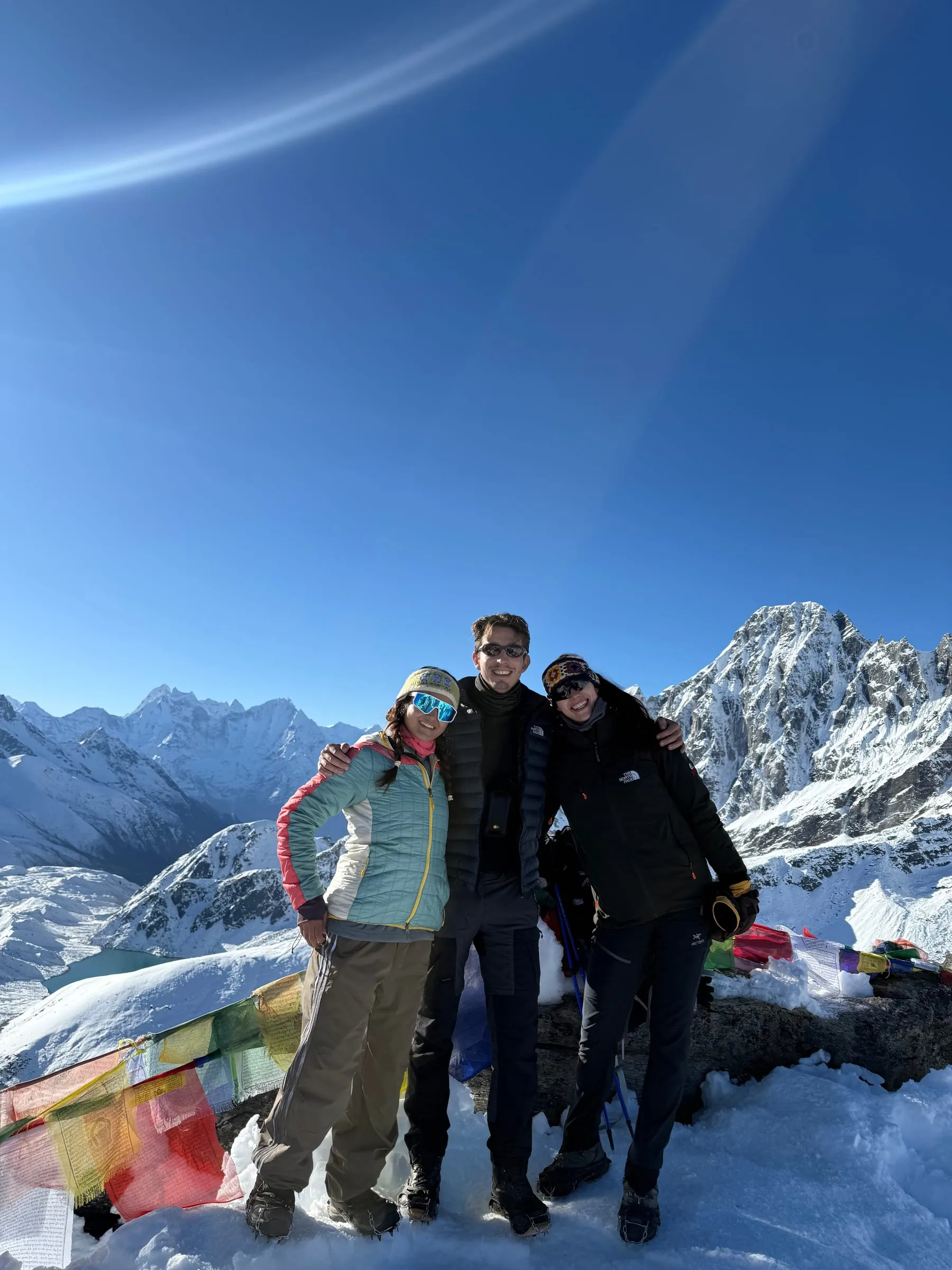 Three trekkers standing arm in arm high on a snowy ridge above the Gokyo valley, prayer flags at their feet and a wall of white peaks behind them.