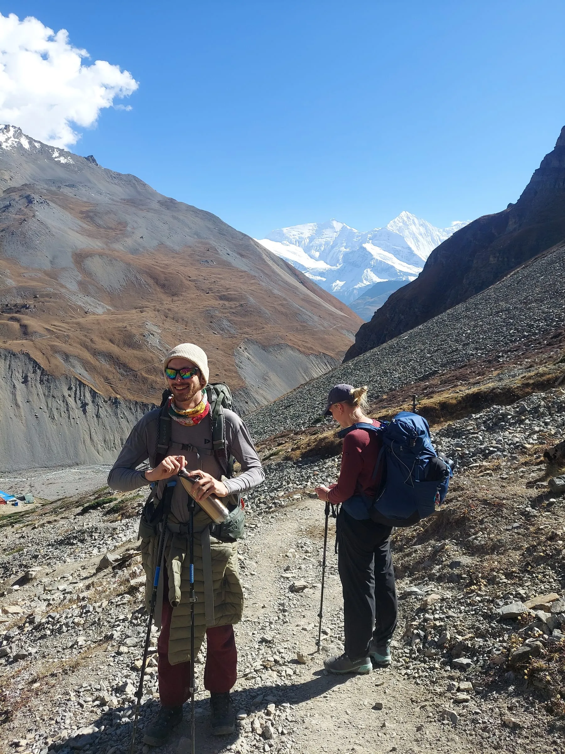 A guide leading the way along a high valley trail, a trekker following behind and snow peaks at the head of the valley.