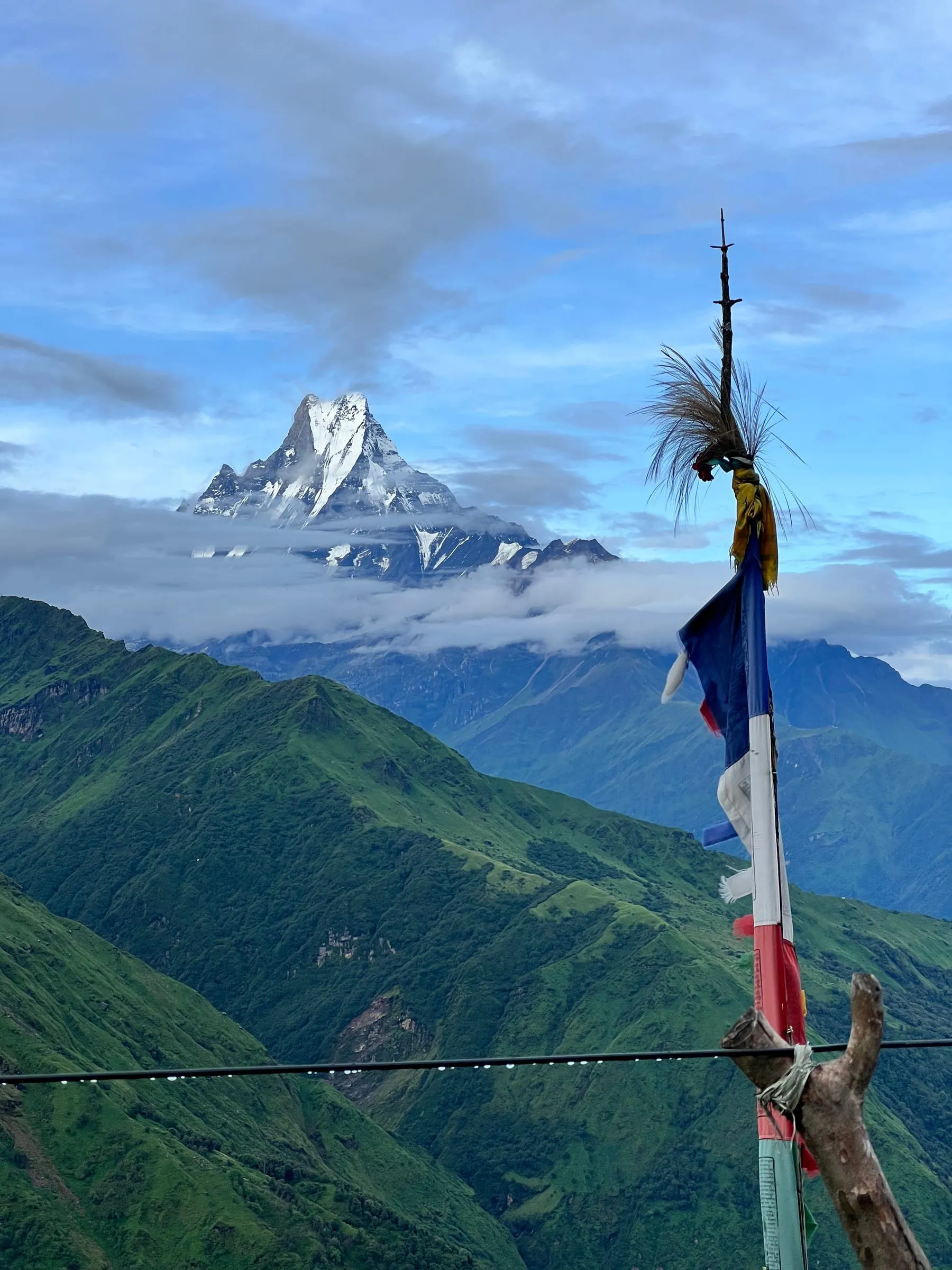Machhapuchhre rising out of cloud above green foothills, a line of prayer flags on a pole in the foreground.