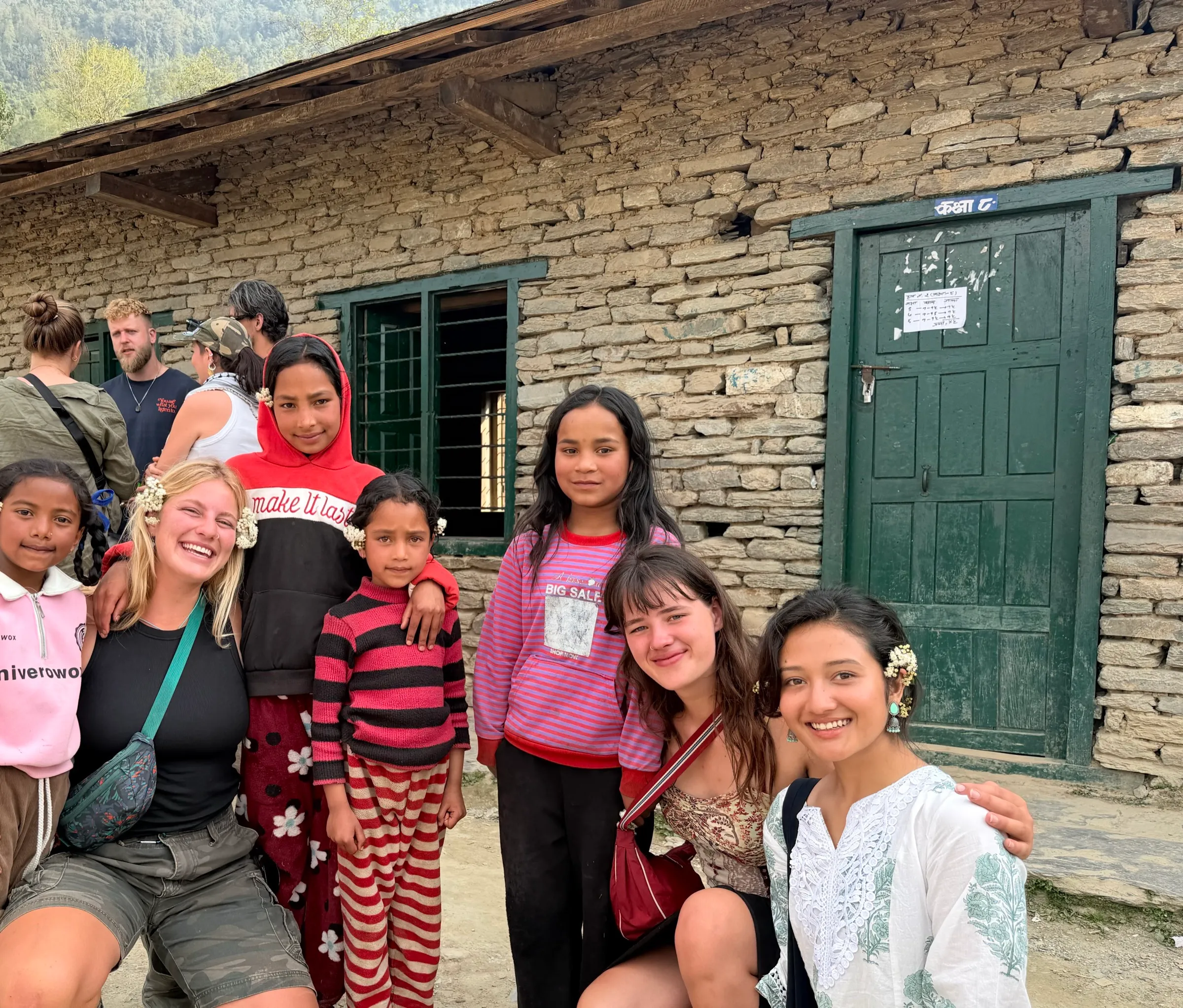 Village children and visitors crouched together for a photograph outside a stone schoolhouse at a village festival, sprigs of white flowers tucked behind their ears.