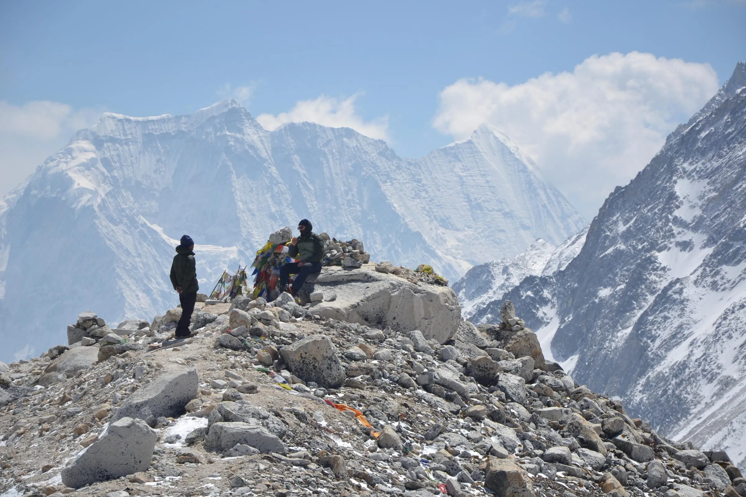 Two figures resting on a rocky ridge beside a cairn strung with prayer flags, a wall of snow peaks across the valley.